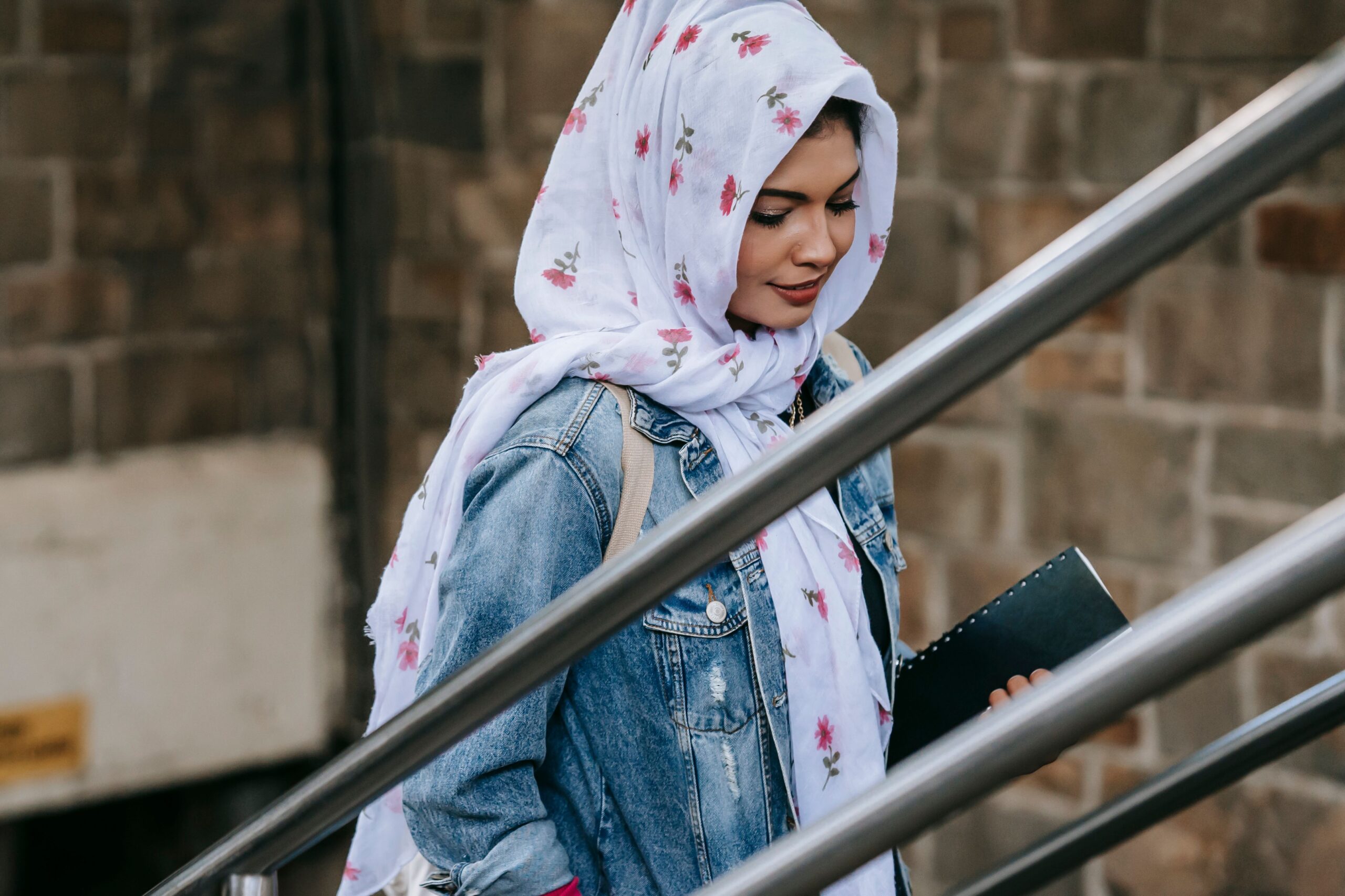 Delighted young Muslim lady wearing denim jacket and headscarf walking upstairs near metal railing from underpass in daytime
